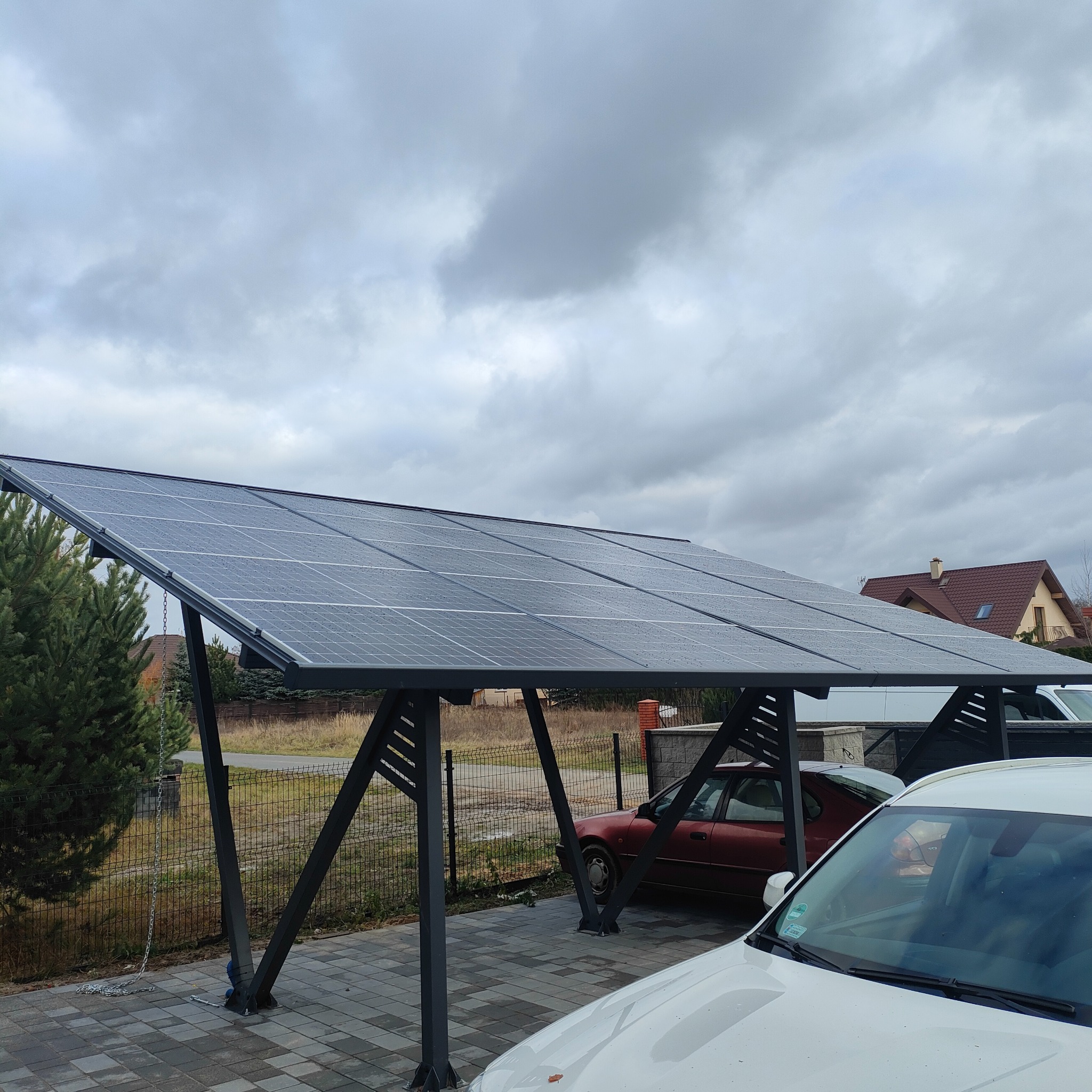 Zadaszenie carport z panelami fotowoltaicznymi w deszczowy dzień, widoczne dwa samochody pod konstrukcją.