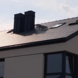 Nowoczesny dom z dachem pokrytym brązową dachówką ceramiczną, widoczne dwa czarne kominy, okna dachowe oraz system przeciwśniegowy.