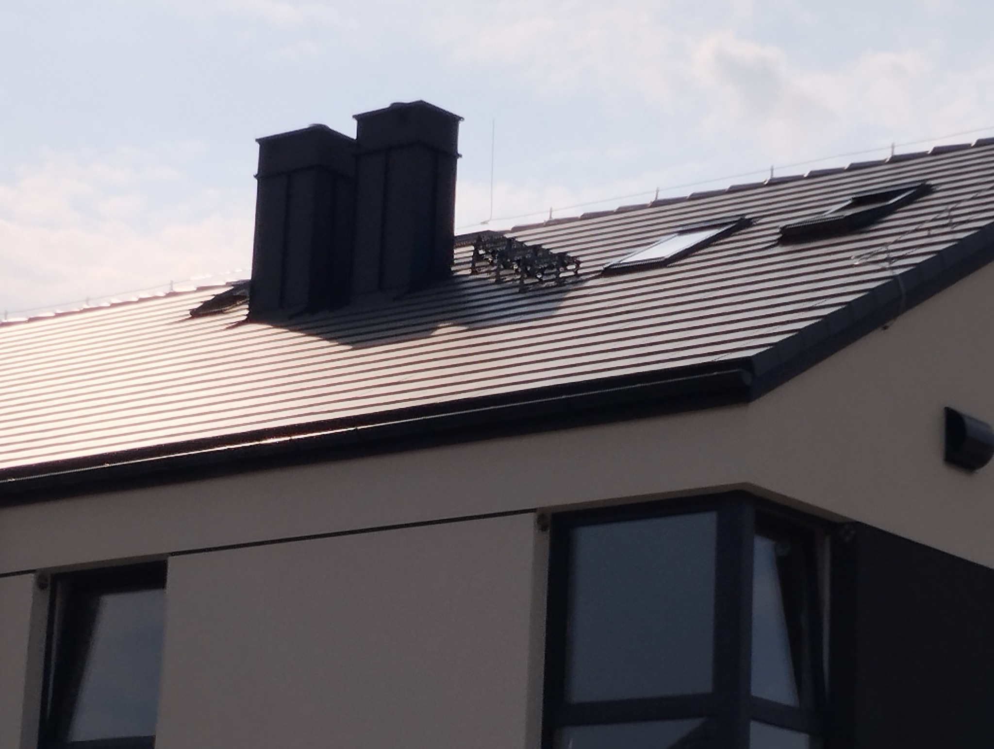 Nowoczesny dom z dachem pokrytym brązową dachówką ceramiczną, widoczne dwa czarne kominy, okna dachowe oraz system przeciwśniegowy.