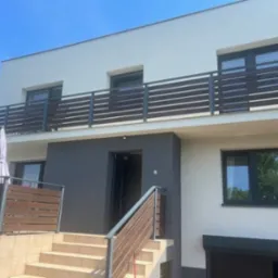 Dwupiętrowy dom z jasną elewacją, ciemnoszarym akcentem przy wejściu, balkonem z drewnianą balustradą i schodami prowadzącymi do wejścia.