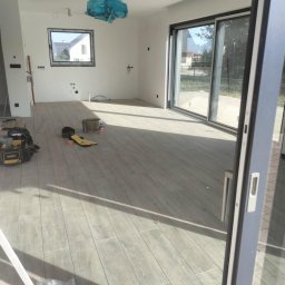 Marcin Kowalczyk BUD - Salon w trakcie prac wykończeniowych: ułożona podłoga z płytek imitujących drewno, narzędzia pracy i akcesoria budowlane rozłożone na podłodze, okno i przesuwne drzwi balkonowe z widokiem...