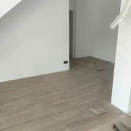 Układanie podłogi z płytek imitujących drewno w salonie, widoczne płytki dystansowe i zapasowe płytki przy ścianie, w tle schody i surowa ściana z otwartym przejściem.