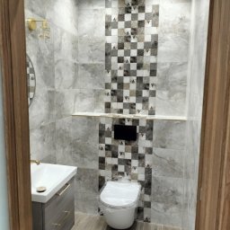 Marcin Kowalczyk BUD - Wykończona łazienka z szarymi płytkami imitującymi marmur, dekoracyjnym pasem płytek w szachownicę, podwieszaną umywalką z szufladami w kolorze grafitowym i złotymi uchwytami, oraz toaletą owiniętą...