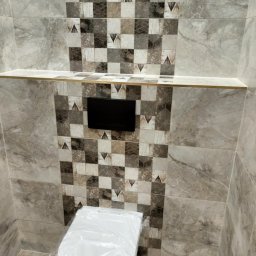 Marcin Kowalczyk BUD - Wykończona łazienka z szarą mozaiką na ścianie, podwieszana toaleta zabezpieczona folią, półka z wykończeniem w kolorze złotym oraz czarny panel sterujący spłuczką.