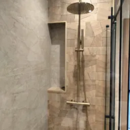 Nowoczesny prysznic z deszczownicą w odcieniach beżu i złota, z geometryczną mozaiką na ścianie i wnęką w marmurowej płytce.