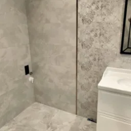 Wykończona łazienka z beżowymi płytkami imitującymi marmur, wzorzysta ściana akcentująca, biała szafka umywalkowa z czarną ramą lustra w tle.