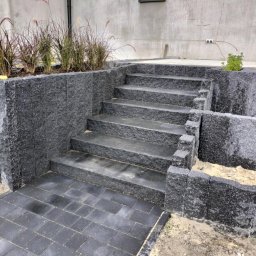 Marcin Kowalczyk BUD - Wykonany z ciemnego kamienia taras z schodami prowadzącymi do ogrodu, obok rabata z trawami ozdobnymi, widoczne niedociągnięcia w postaci piasku na kostce i ziemi przy obrzeżach.