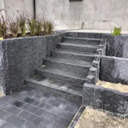 Wykonany z ciemnego kamienia taras z schodami prowadzącymi do ogrodu, obok rabata z trawami ozdobnymi, widoczne niedociągnięcia w postaci piasku na kostce i ziemi przy obrzeżach.