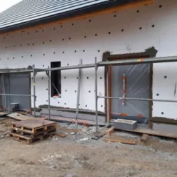 Dom w trakcie ocieplania: białe płyty styropianowe z czarnymi kołkami, częściowo pokryte szarą warstwą kleju, zabezpieczone folią okna i drzwi, rusztowanie, palety, bałagan na ziemi.
