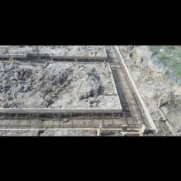 Przygotowanie zbrojenia i szalunku pod wylewkę fundamentów, widoczne drewniane stemple i stalowe pręty, ziemia częściowo rozkopana.