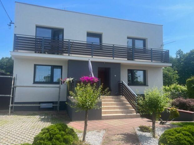 Dwupiętrowy dom z białymi ścianami i ciemnoszarą wnęką wejściową, balkon z poziomymi barierkami, widoczne rusztowanie po lewej stronie, zadbany ogród z niskimi krzewami i drzewkami.