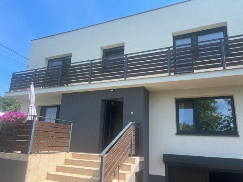 Dwupiętrowy dom z jasną elewacją, ciemnoszarym akcentem przy wejściu, balkonem z drewnianą balustradą i schodami prowadzącymi do wejścia.