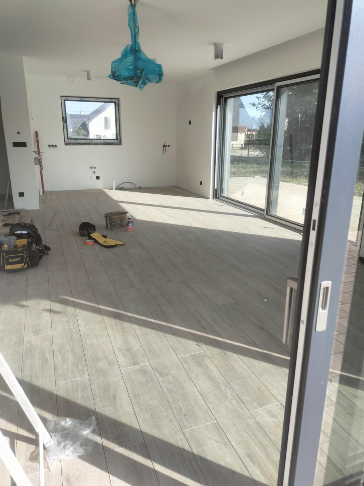 Salon w trakcie prac wykończeniowych: ułożona podłoga z płytek imitujących drewno, narzędzia pracy i akcesoria budowlane rozłożone na podłodze, okno i przesuwne drzwi balkonowe z widokiem...