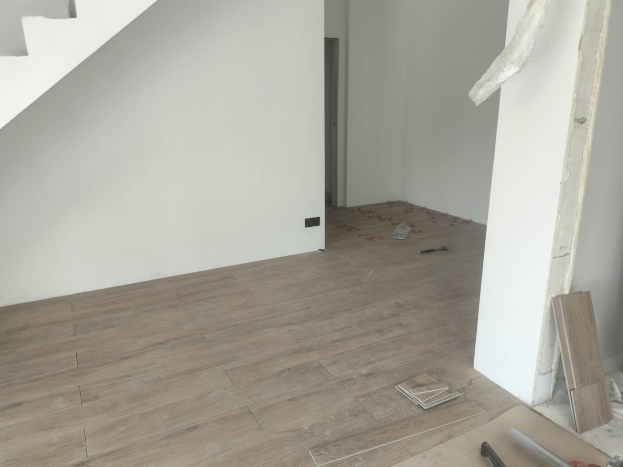 Układanie podłogi z płytek imitujących drewno w salonie, widoczne płytki dystansowe i zapasowe płytki przy ścianie, w tle schody i surowa ściana z otwartym przejściem.