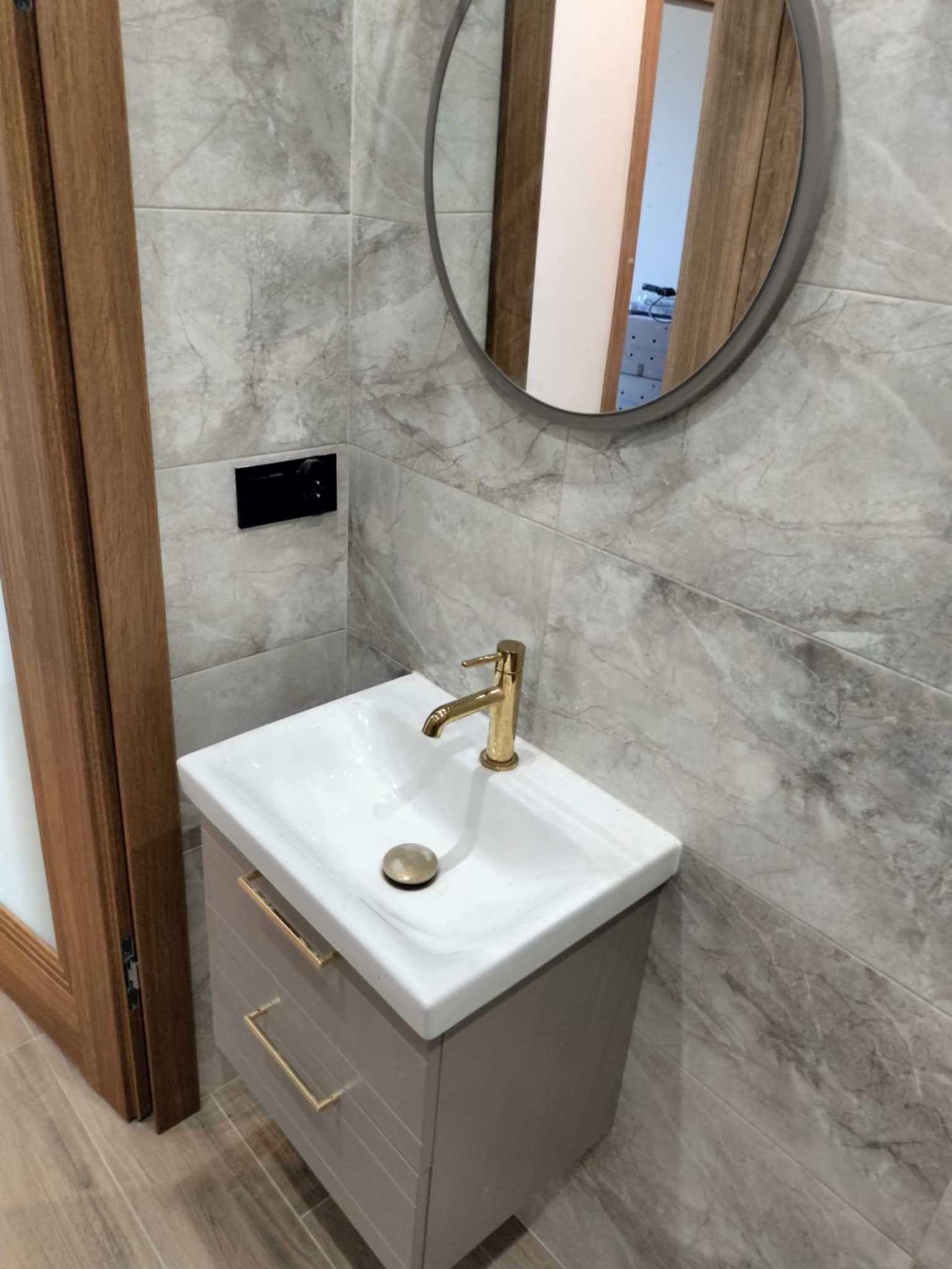 Nowoczesna łazienka z umywalką nablatową, złotą armaturą i lustrem w okrągłej ramie, płytki imitujące marmur na ścianach i podłodze, widoczny fragment drewnianej ościeżnicy drzwi.