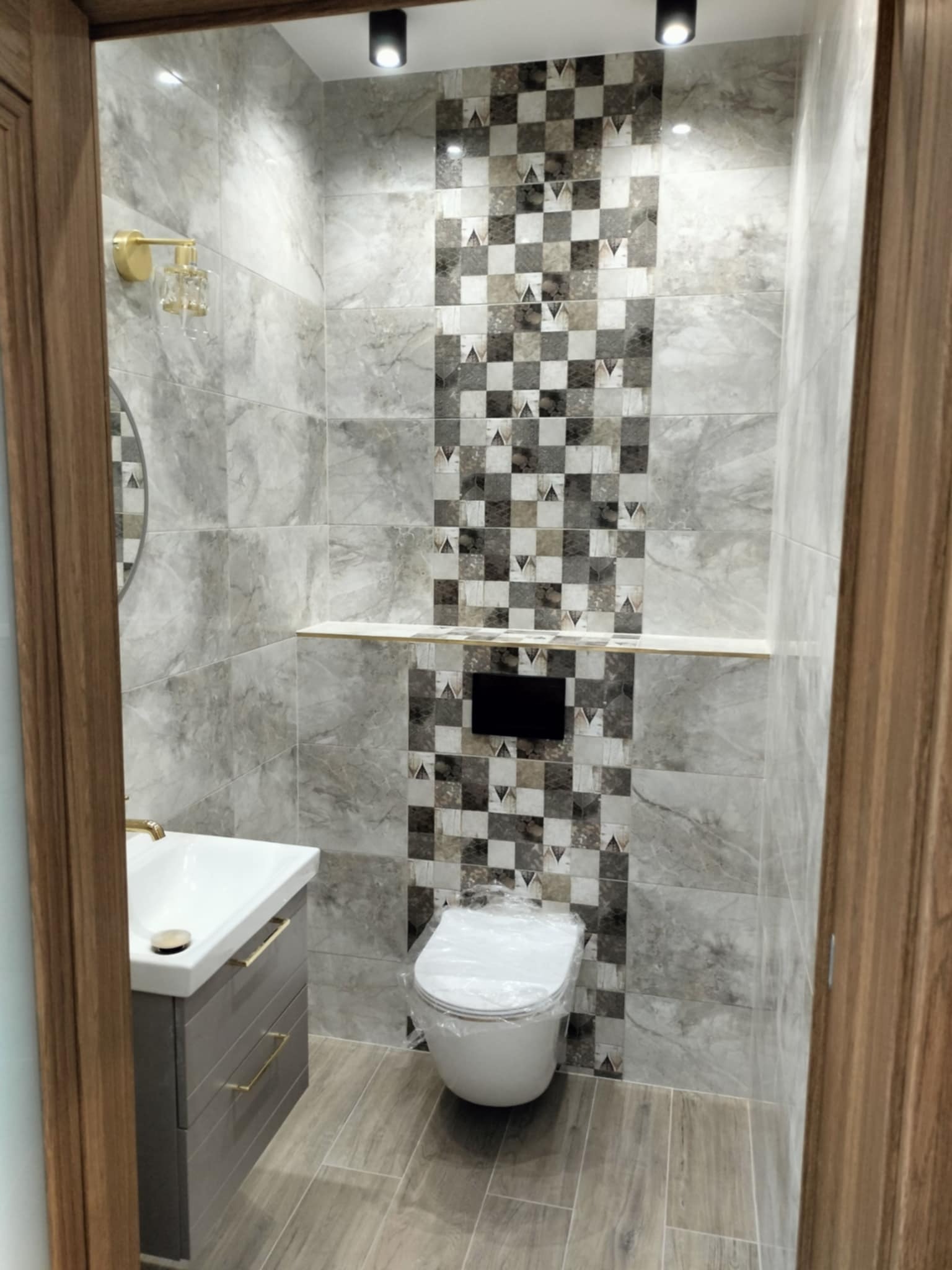 Wykończona łazienka z szarymi płytkami imitującymi marmur, dekoracyjnym pasem płytek w szachownicę, podwieszaną umywalką z szufladami w kolorze grafitowym i złotymi uchwytami, oraz toaletą owiniętą...