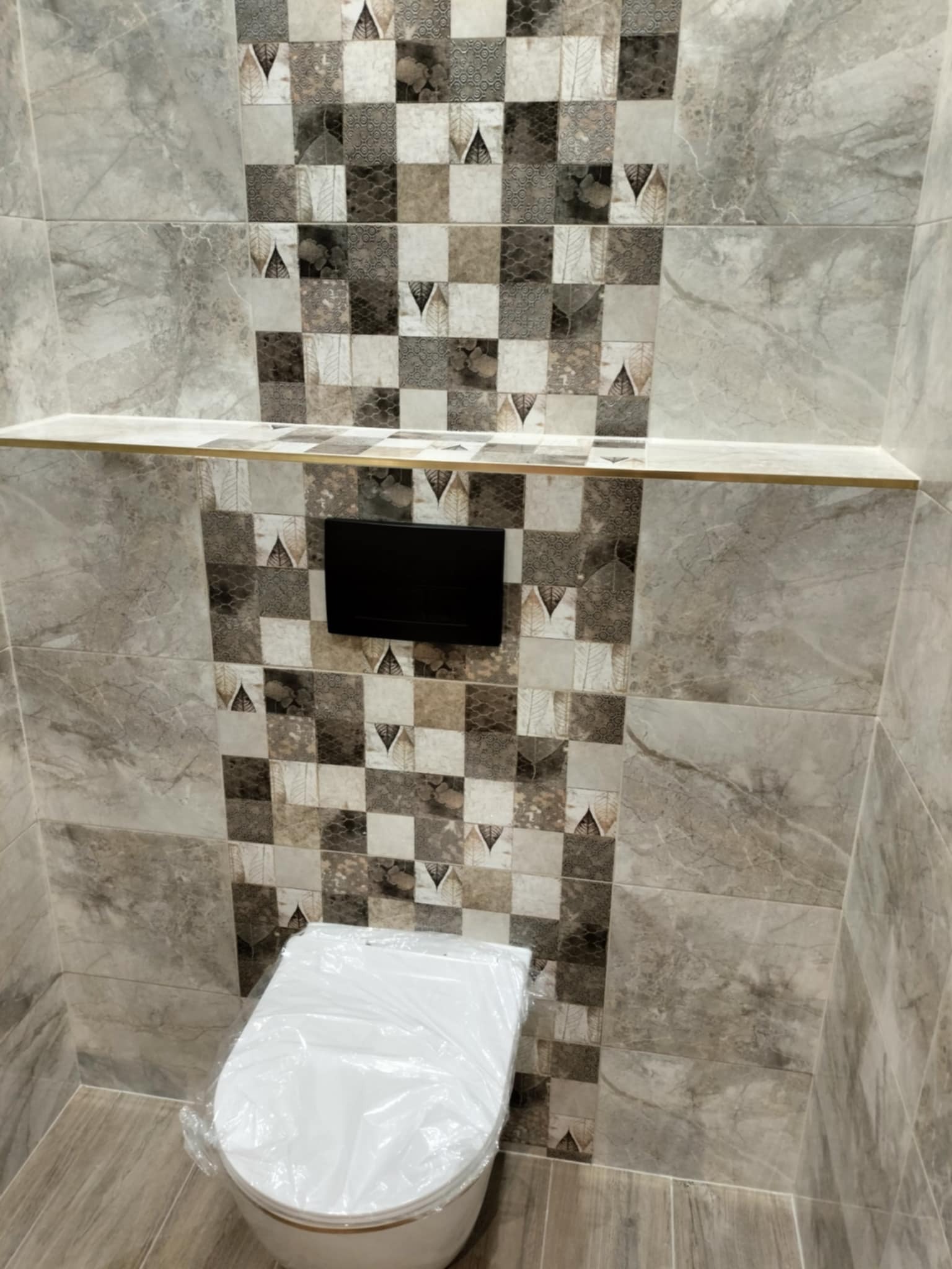 Wykończona łazienka z szarą mozaiką na ścianie, podwieszana toaleta zabezpieczona folią, półka z wykończeniem w kolorze złotym oraz czarny panel sterujący spłuczką.