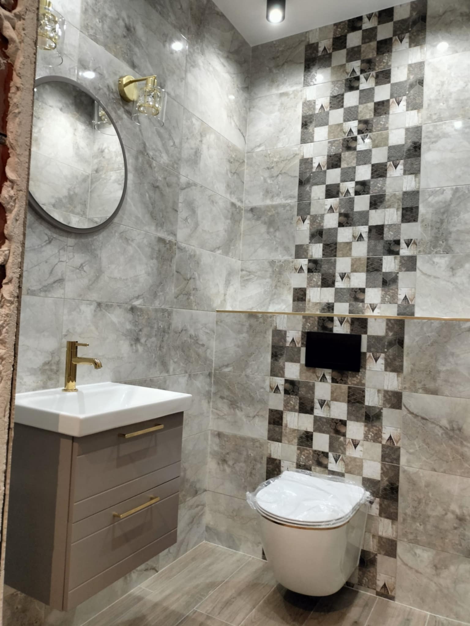 Wykończona łazienka z marmurowymi płytkami, dekoracyjnym pasem mozaiki, wiszącą szafką z umywalką i złotą armaturą oraz toaletą z folią ochronną na desce.