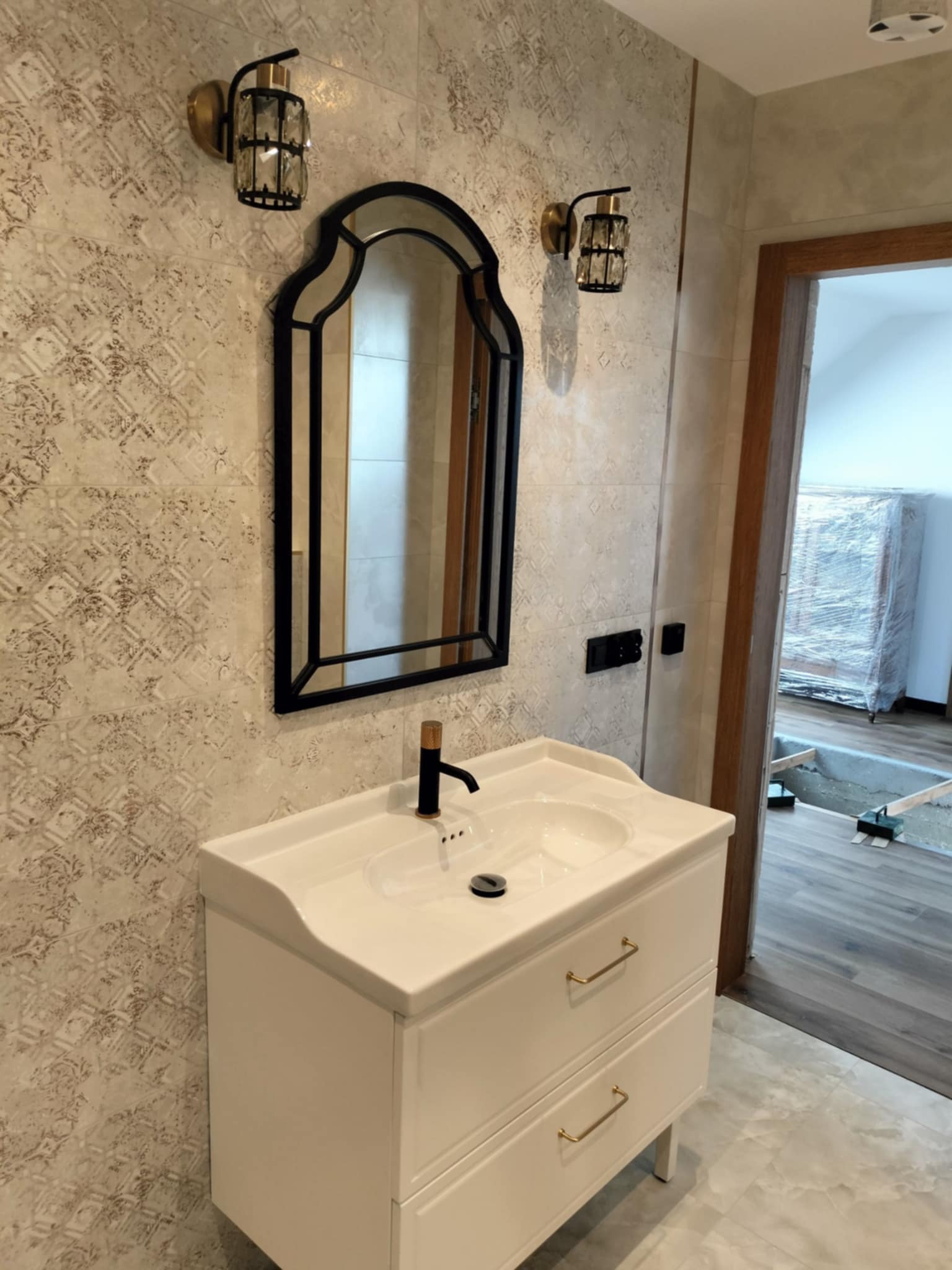 Umywalka z szafką w stylu glamour, czarna armatura, ozdobne kinkiety i lustro w czarnej ramie na tle wzorzystych płytek, w tle widoczny pokój w trakcie wykończenia.