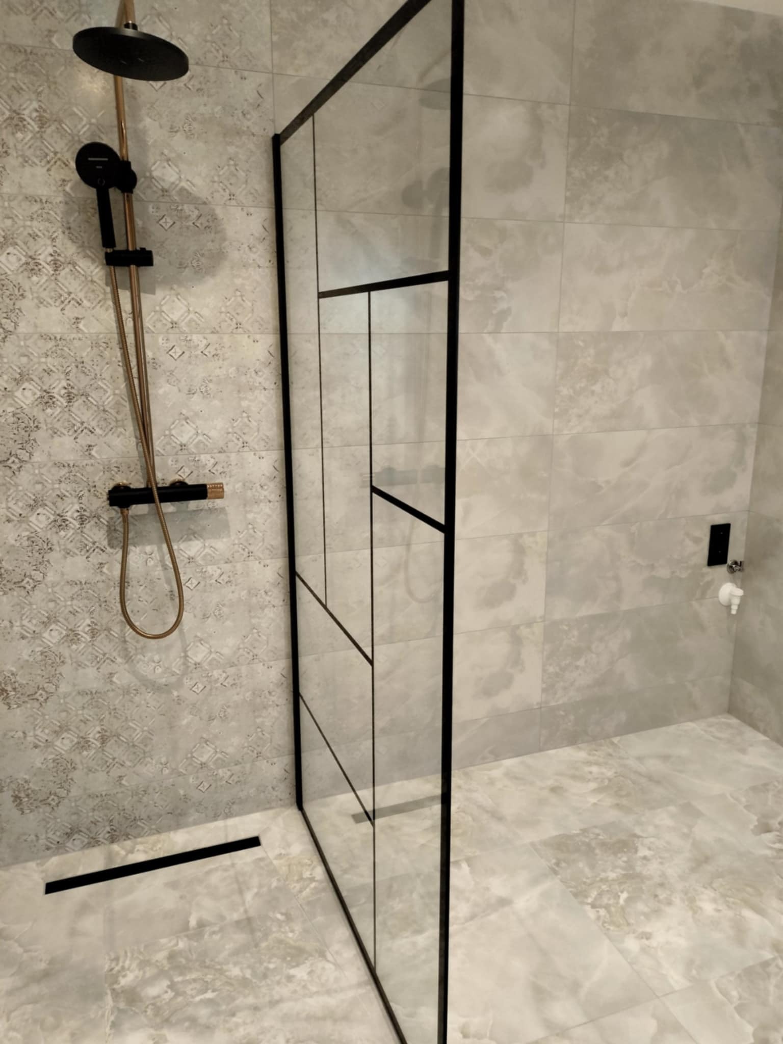 Nowoczesna kabina prysznicowa walk-in z czarnymi profilami i odpływem liniowym, szare płytki imitujące marmur na ścianach i podłodze, bateria prysznicowa w kolorze czarnym i miedzianym.