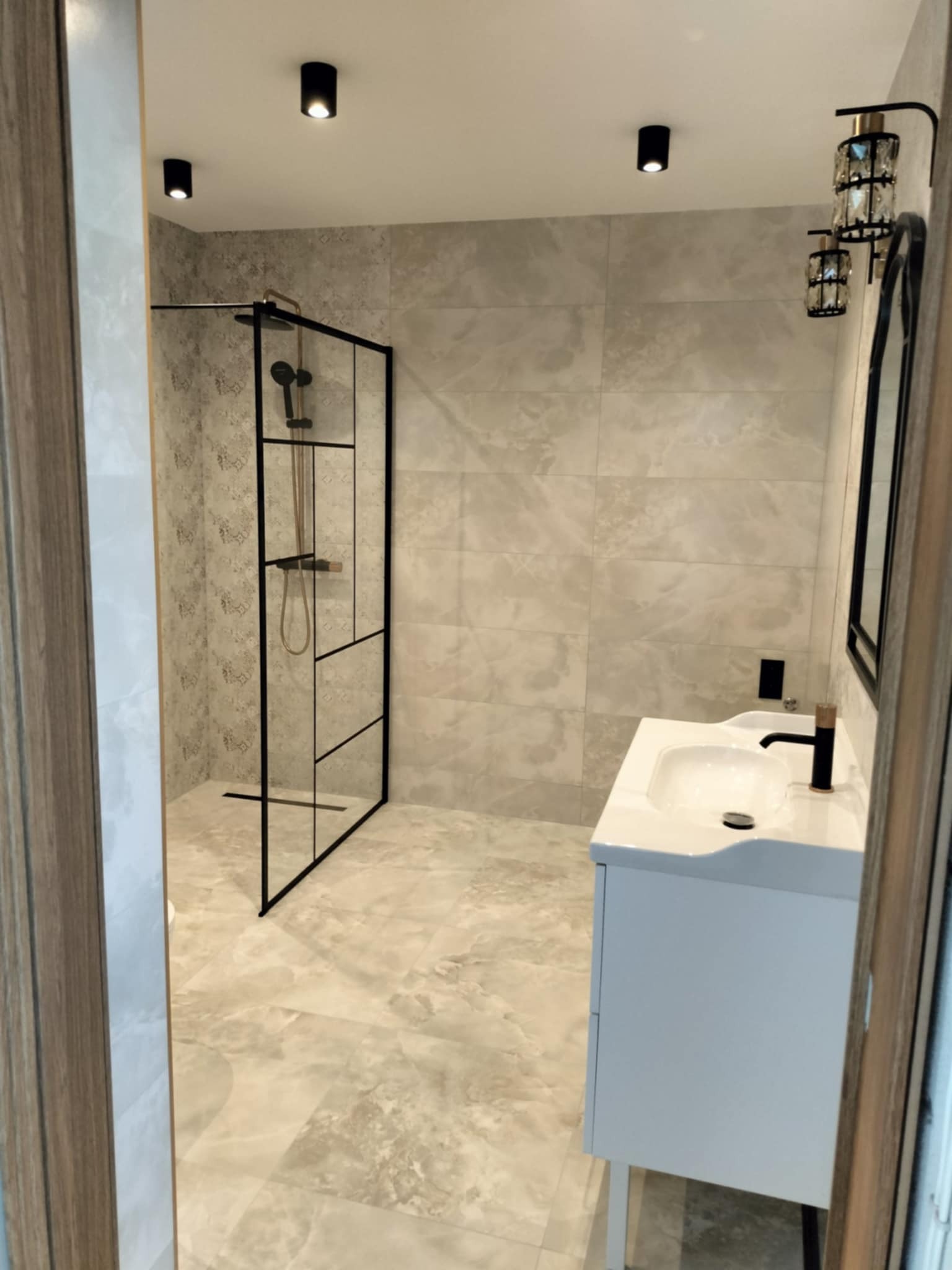 Nowoczesna łazienka w odcieniach beżu i szarości z kabiną prysznicową typu walk-in z czarnymi profilami, białą szafką z umywalką i czarną armaturą oraz dekoracyjnymi lampami kryształowymi.