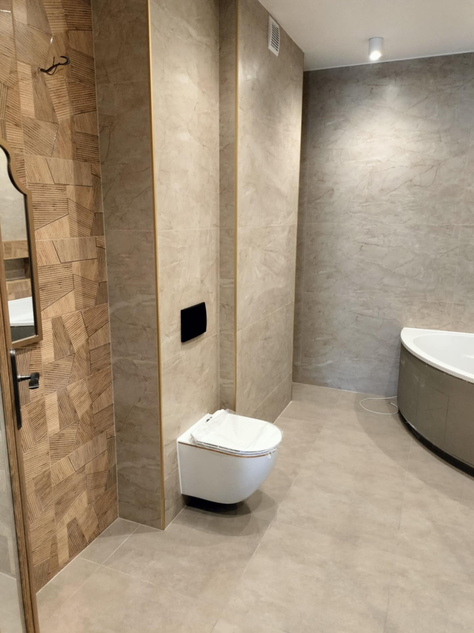 Nowoczesna łazienka z beżowymi płytkami imitującymi marmur, wisząca toaleta, dekoracyjna ściana z drewnopodobnych płytek i fragment wanny.