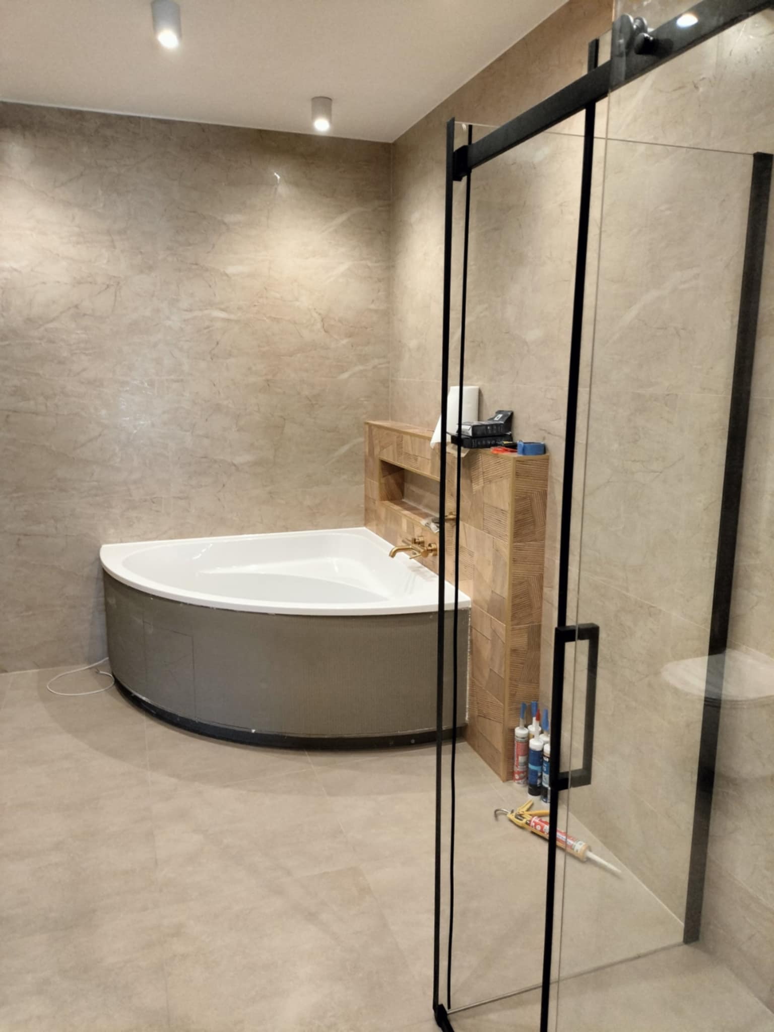 Nowoczesna łazienka z narożną wanną, obudowaną drewnopodobną mozaiką, kabiną prysznicową z czarnymi profilami i beżowymi płytkami imitującymi marmur na ścianach i podłodze; widoczne silikony...