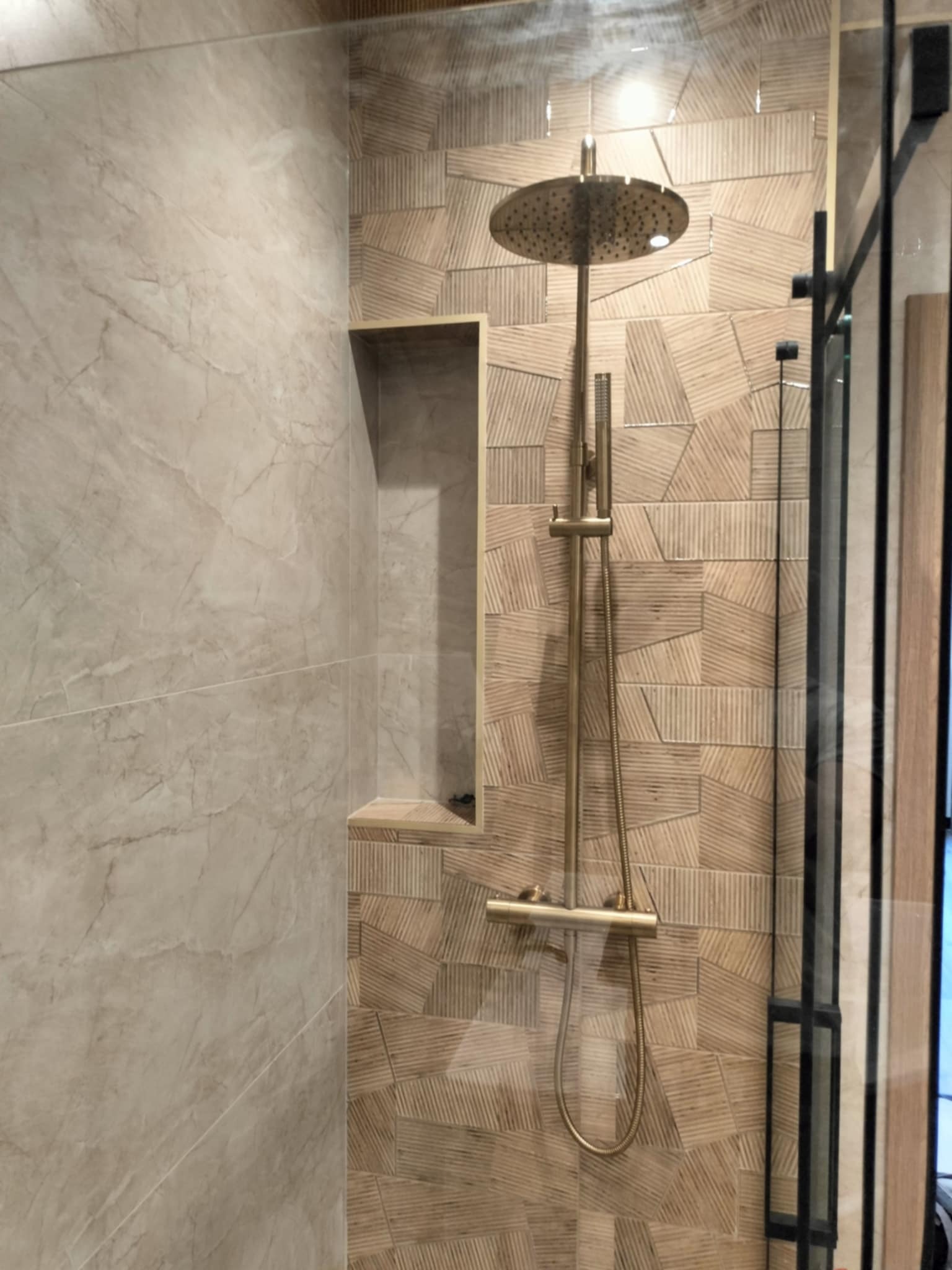 Nowoczesny prysznic z deszczownicą w odcieniach beżu i złota, z geometryczną mozaiką na ścianie i wnęką w marmurowej płytce.