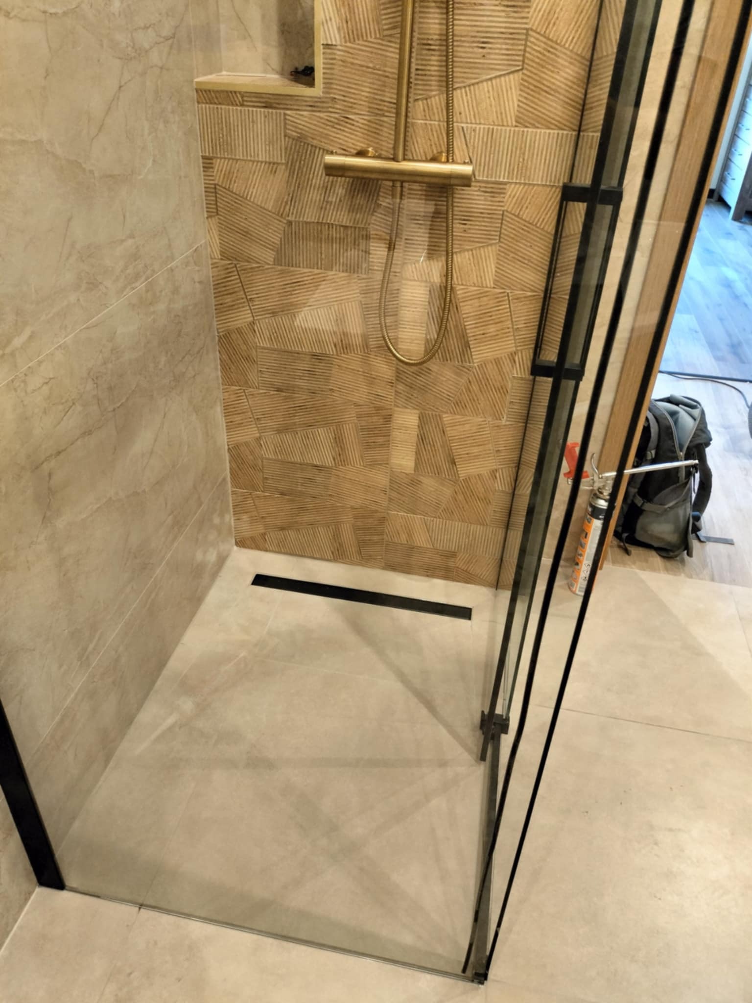 Nowoczesna kabina prysznicowa walk-in z czarnymi profilami, złotą armaturą i odpływem liniowym, wykończona beżowymi płytkami o zróżnicowanej fakturze. W tle widoczny plecak oraz pistolet do pianki...