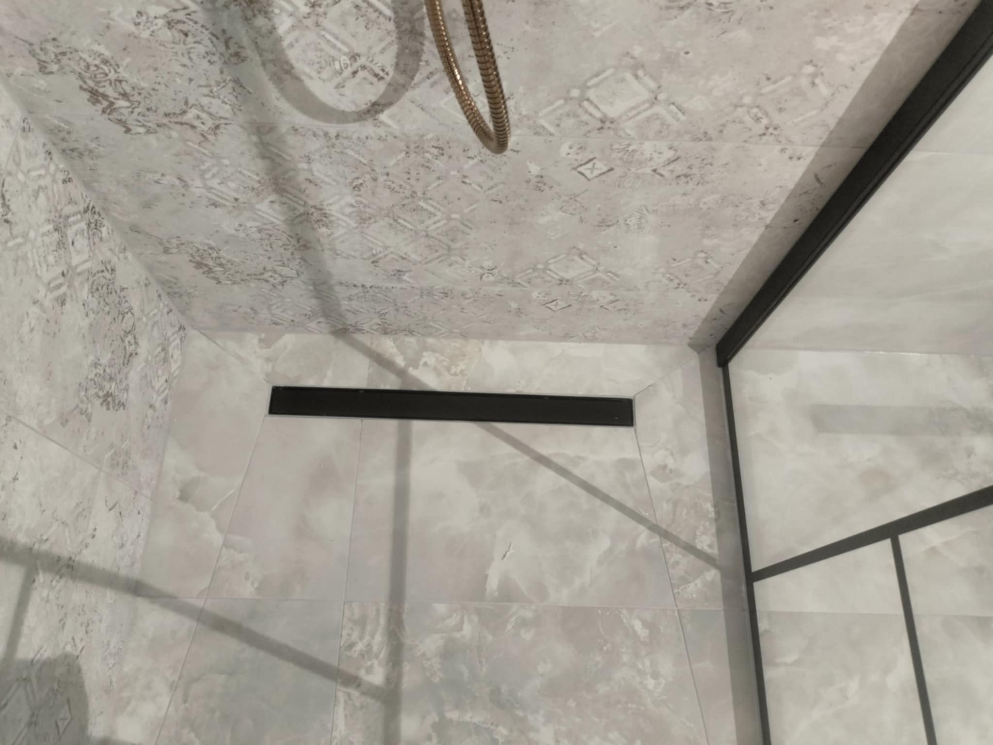 Wykończona strefa prysznicowa z odpływem liniowym, wyłożona płytkami imitującymi marmur w odcieniach szarości i bieli, widok z góry na sufit z dekoracyjną fakturą i elementem deszczownicy.