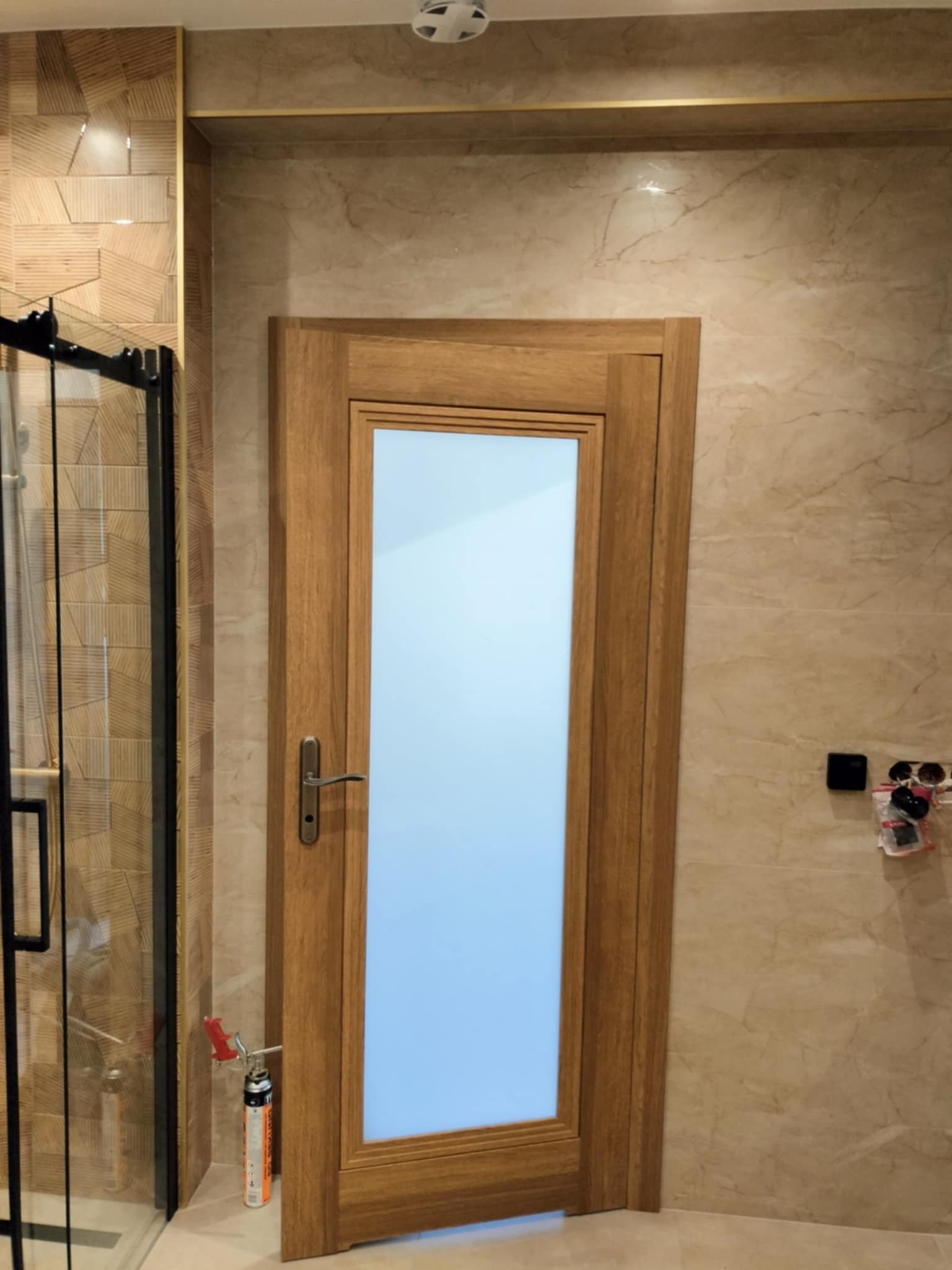 Nowoczesna łazienka z drewnianą ramą drzwi z mlecznym szkłem, beżowe płytki imitujące marmur na ścianach, kabina prysznicowa z czarnymi ramami, pistolet do pianki montażowej obok drzwi.