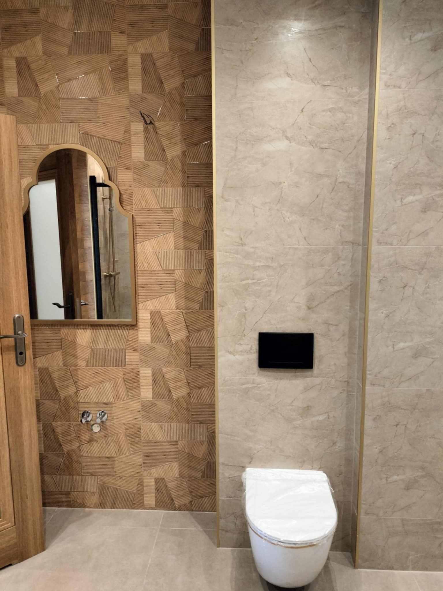 Wykończona łazienka z drewnopodobnymi płytkami ułożonymi w geometryczny wzór na ścianie, lustro w stylu marokańskim, biała toaleta z deską w folii ochronnej i beżowe płytki imitujące marmur...