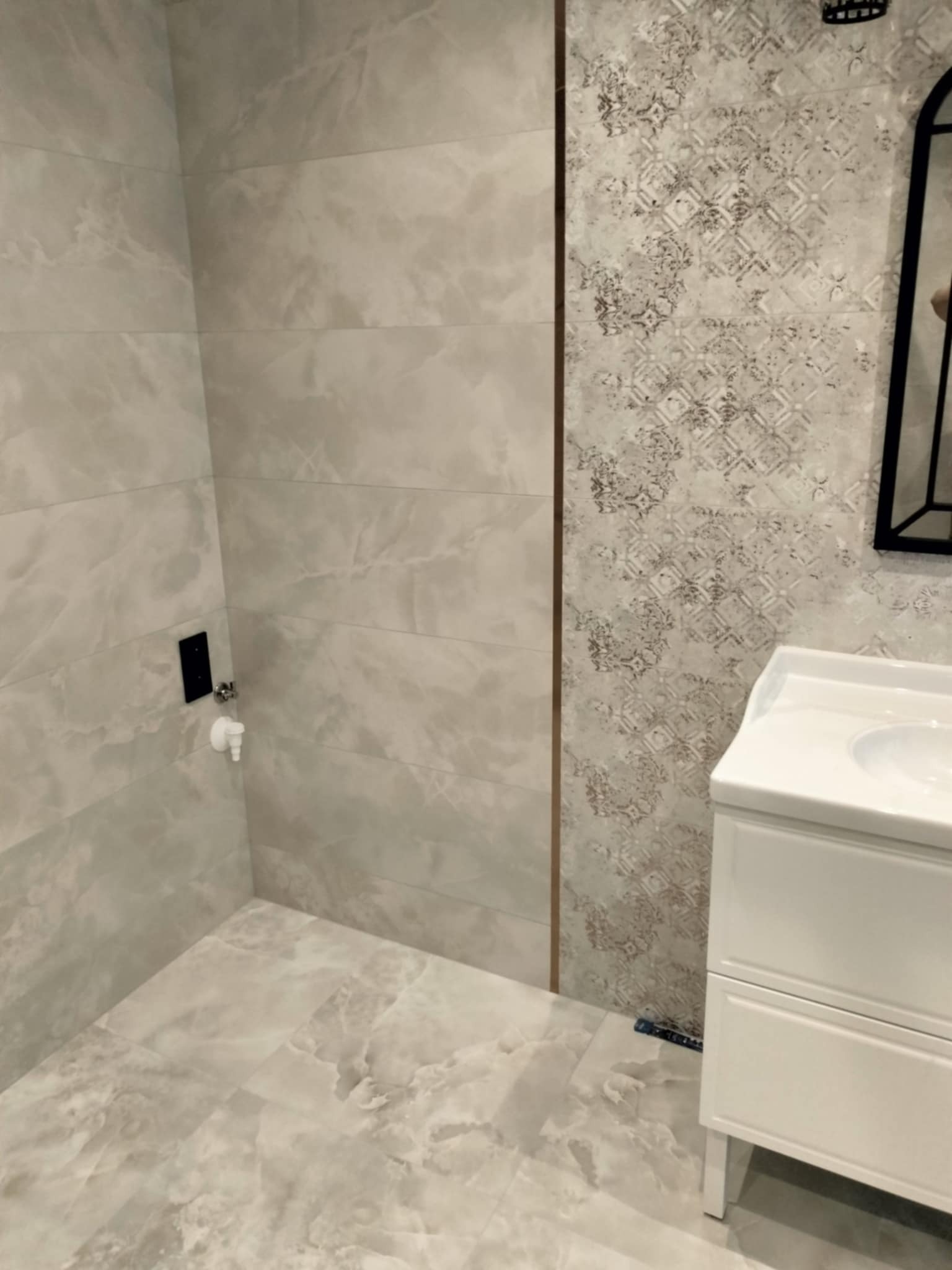 Wykończona łazienka z beżowymi płytkami imitującymi marmur, wzorzysta ściana akcentująca, biała szafka umywalkowa z czarną ramą lustra w tle.