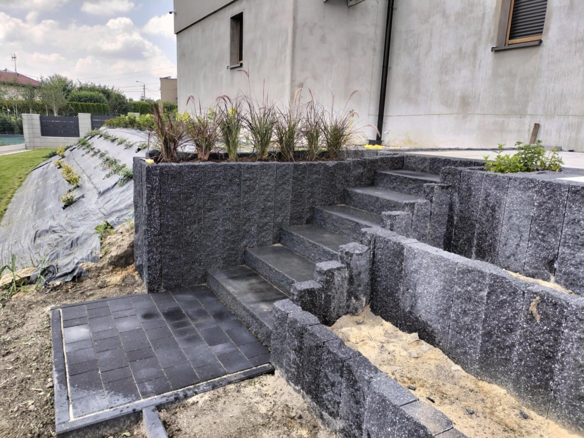Nowo wybudowane, granitowe schody tarasowe prowadzące do domu, z elementami dekoracyjnymi z roślin, w trakcie prac wykończeniowych z widocznym piaskiem i folią ochronną na skarpie.