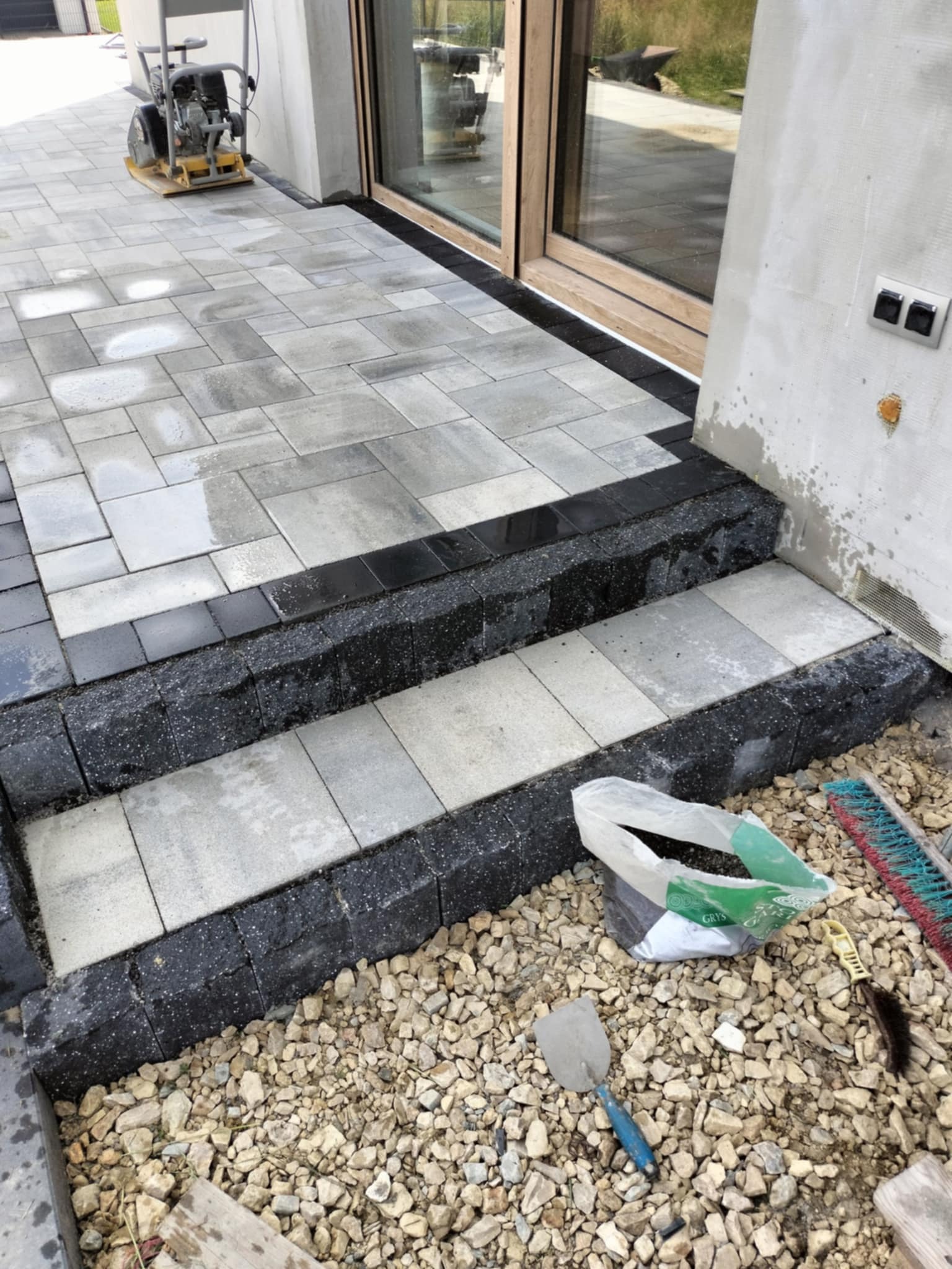 Układanie kostki brukowej na tarasie i schodach wejściowych, widoczne narzędzia, zagęszczarka i częściowo ułożona nawierzchnia z kostki w dwóch kolorach.