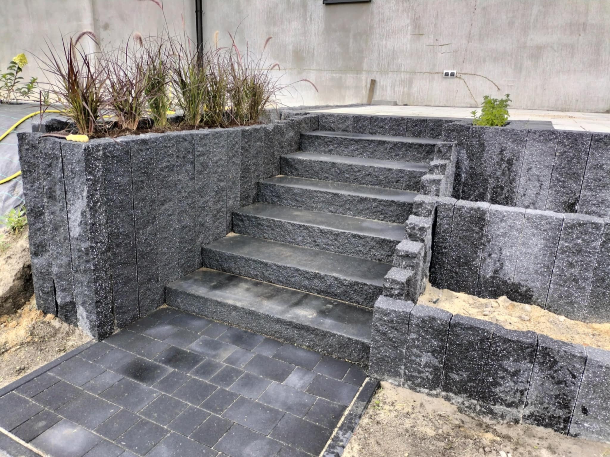 Wykonany z ciemnego kamienia taras z schodami prowadzącymi do ogrodu, obok rabata z trawami ozdobnymi, widoczne niedociągnięcia w postaci piasku na kostce i ziemi przy obrzeżach.