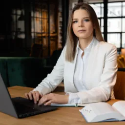 Profesjonalna kobieta w białym garniturze pracuje przy laptopie na drewnianym biurku, otwarty notatnik obok, w tle eleganckie wnętrze z zielonymi i złotymi sofami.