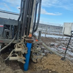 PALABRA JOANNA KLIMASZEWSKA - Wiertnica w trakcie pracy przy studni głębinowej w Skórzcu. Widoczna niebieska rura w ziemi, metalowa krata i sprzęt wiertniczy na tle pochmurnego nieba.