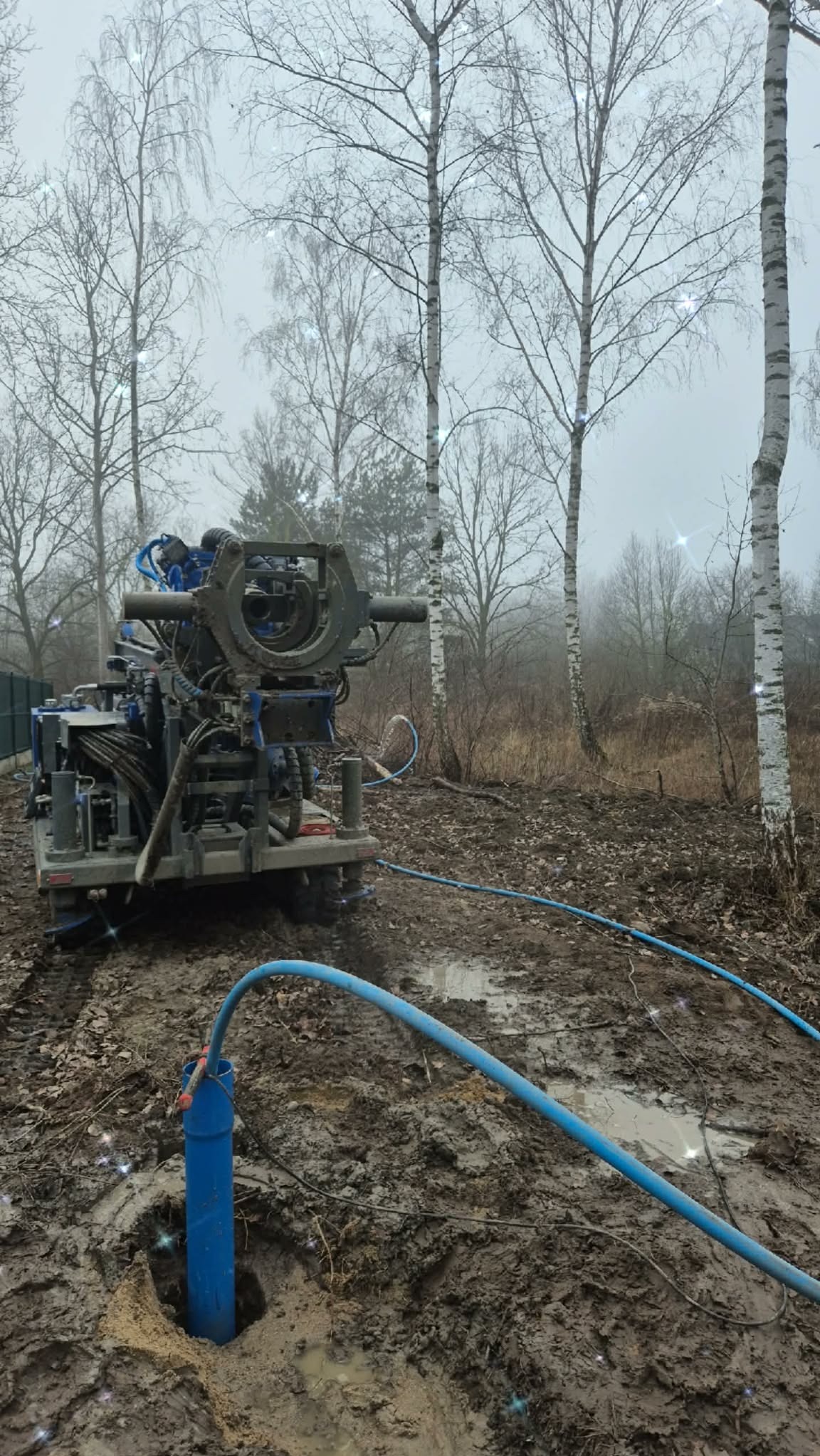 Wiertnica w błotnistym terenie, niebieska rura w ziemi, drzewa w tle. Wykonywanie odwiertu studni głębinowej w pochmurny dzień.