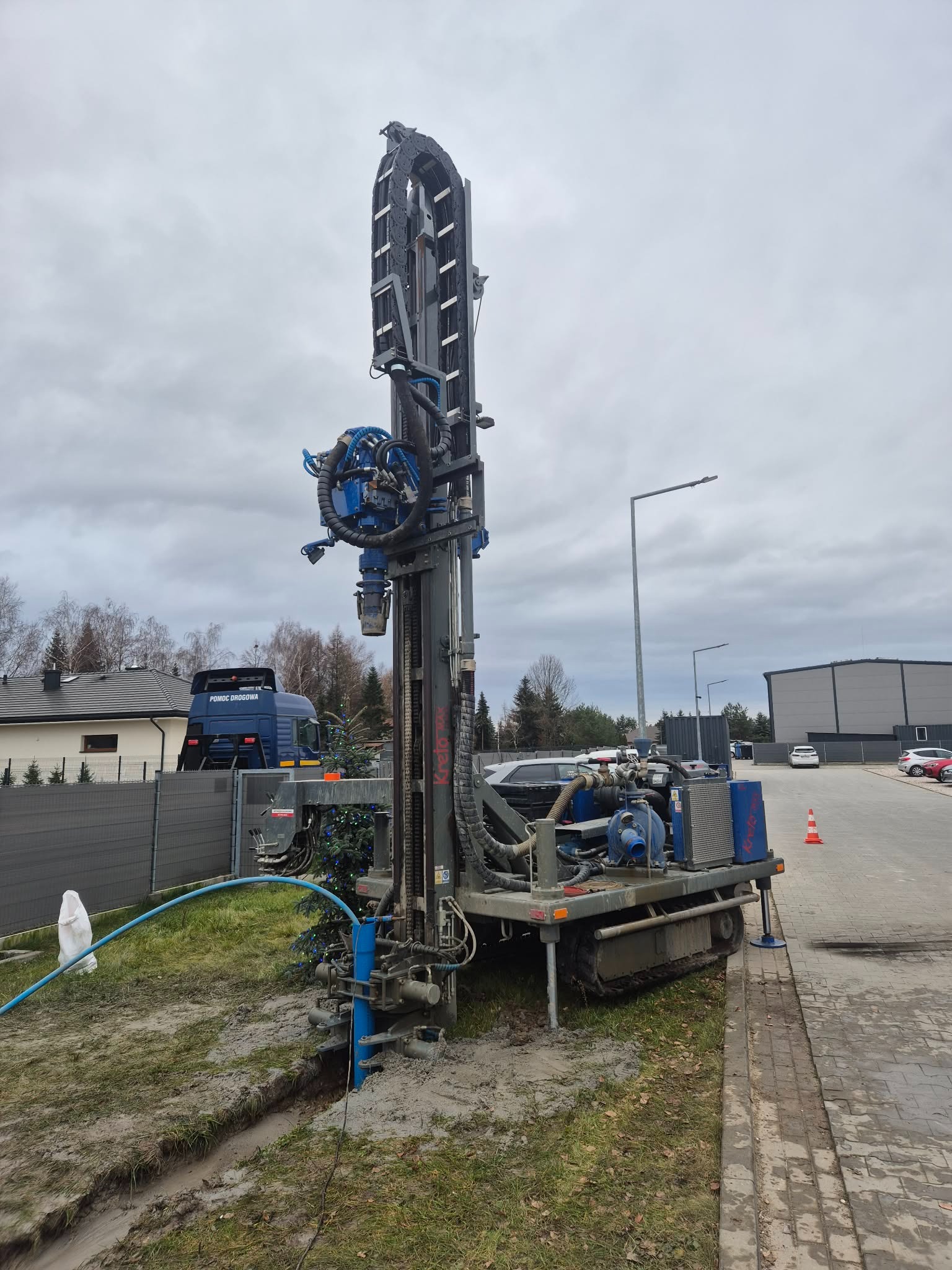 Wiertnica do studni głębinowych na podwoziu gąsienicowym, z niebieskimi elementami, w trakcie pracy na działce. W tle budynki i niebieska ciężarówka pomocy drogowej.
