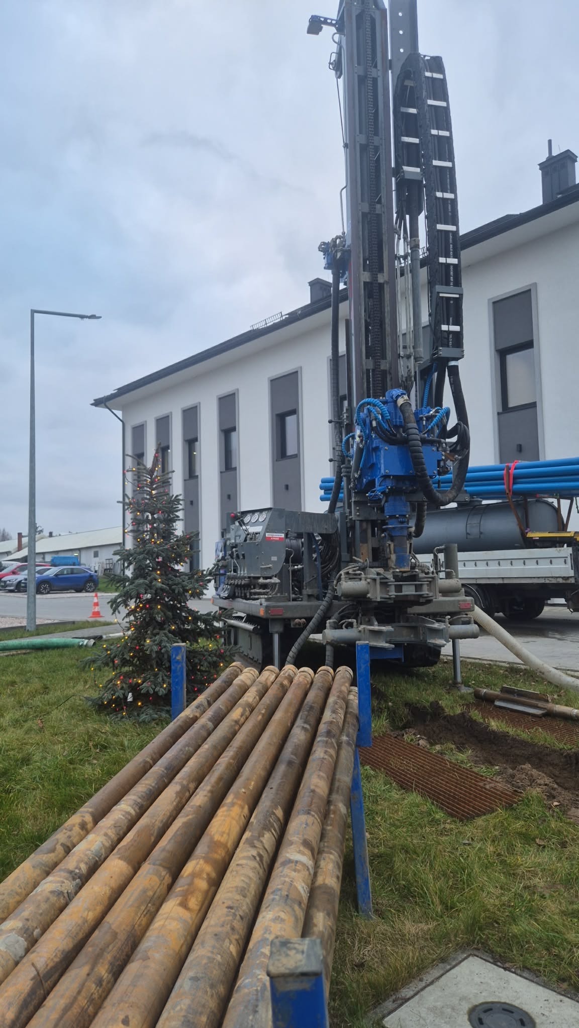Wiertnica do studni głębinowych na tle budynku z choinką, rury wiertnicze na pierwszym planie. Sprzęt gotowy do pracy, widoczne elementy hydrauliczne i konstrukcyjne.