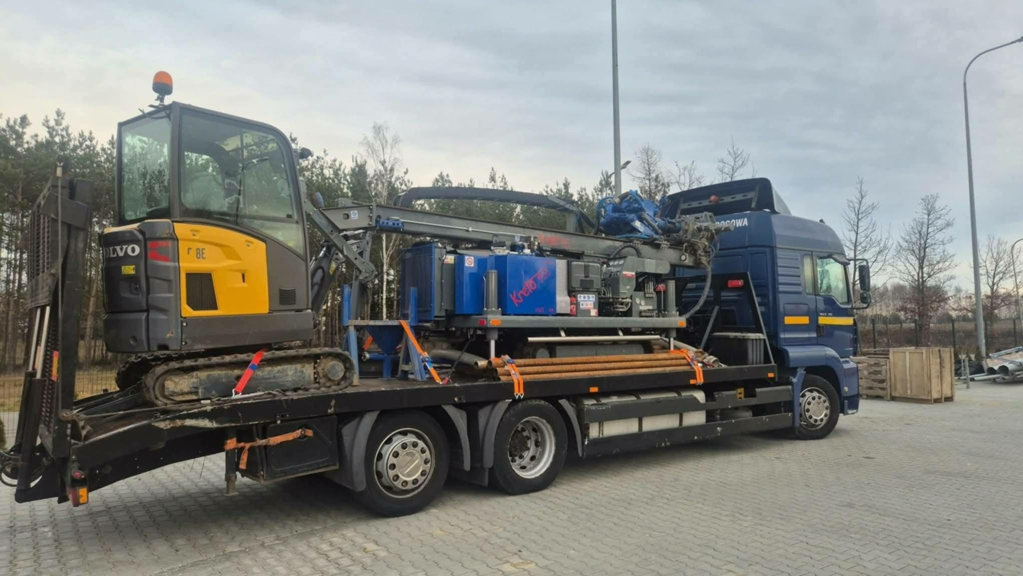 Ciężarówka z platformą wiertniczą i małą koparką Volvo F8E na lawecie. Sprzęt gotowy do transportu na miejsce wiercenia studni głębinowej. Widoczne rury wiertnicze.