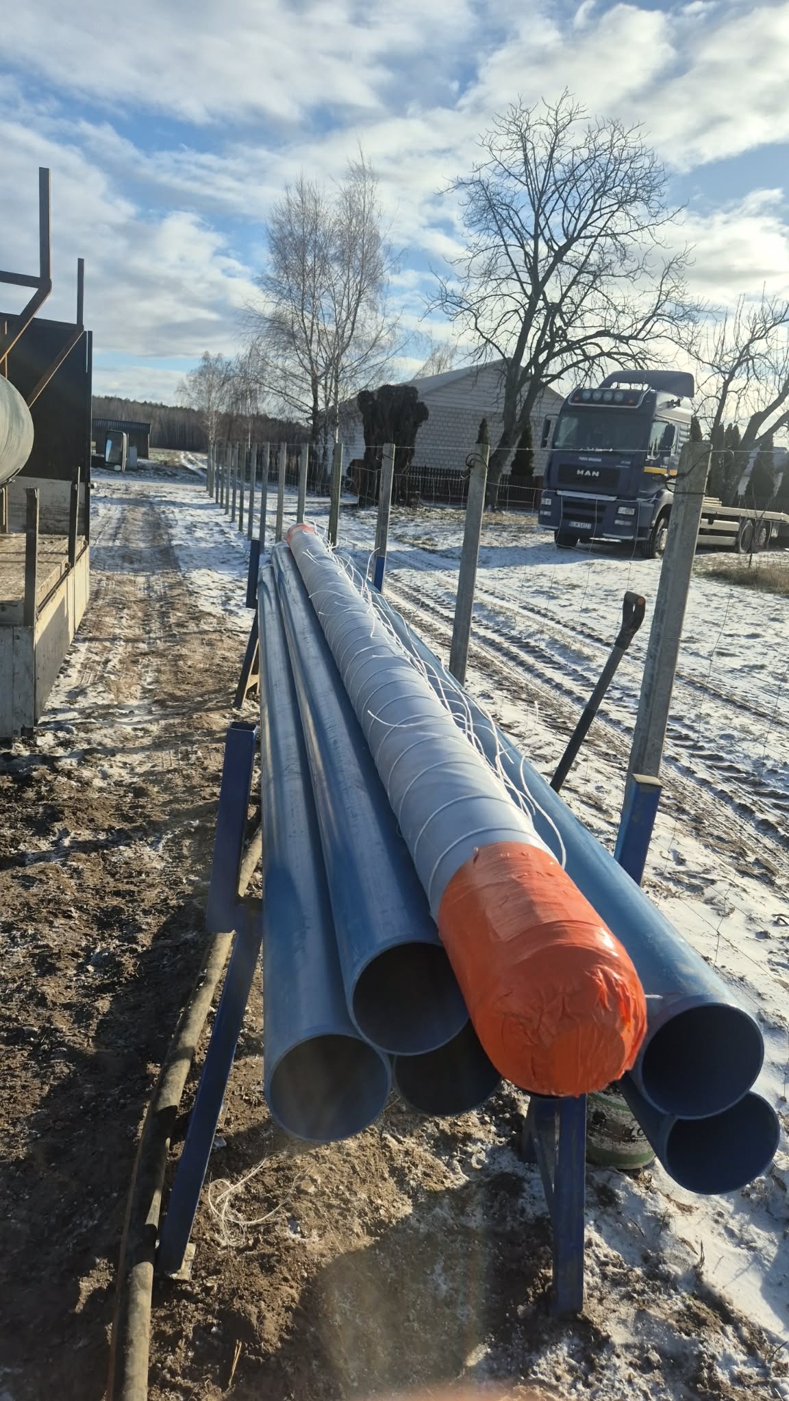 Niebieskie rury na stojaku, zabezpieczone pomarańczową folią, na tle zimowego krajobrazu z widocznym samochodem ciężarowym MAN i ogrodzeniem z drutu kolczastego.