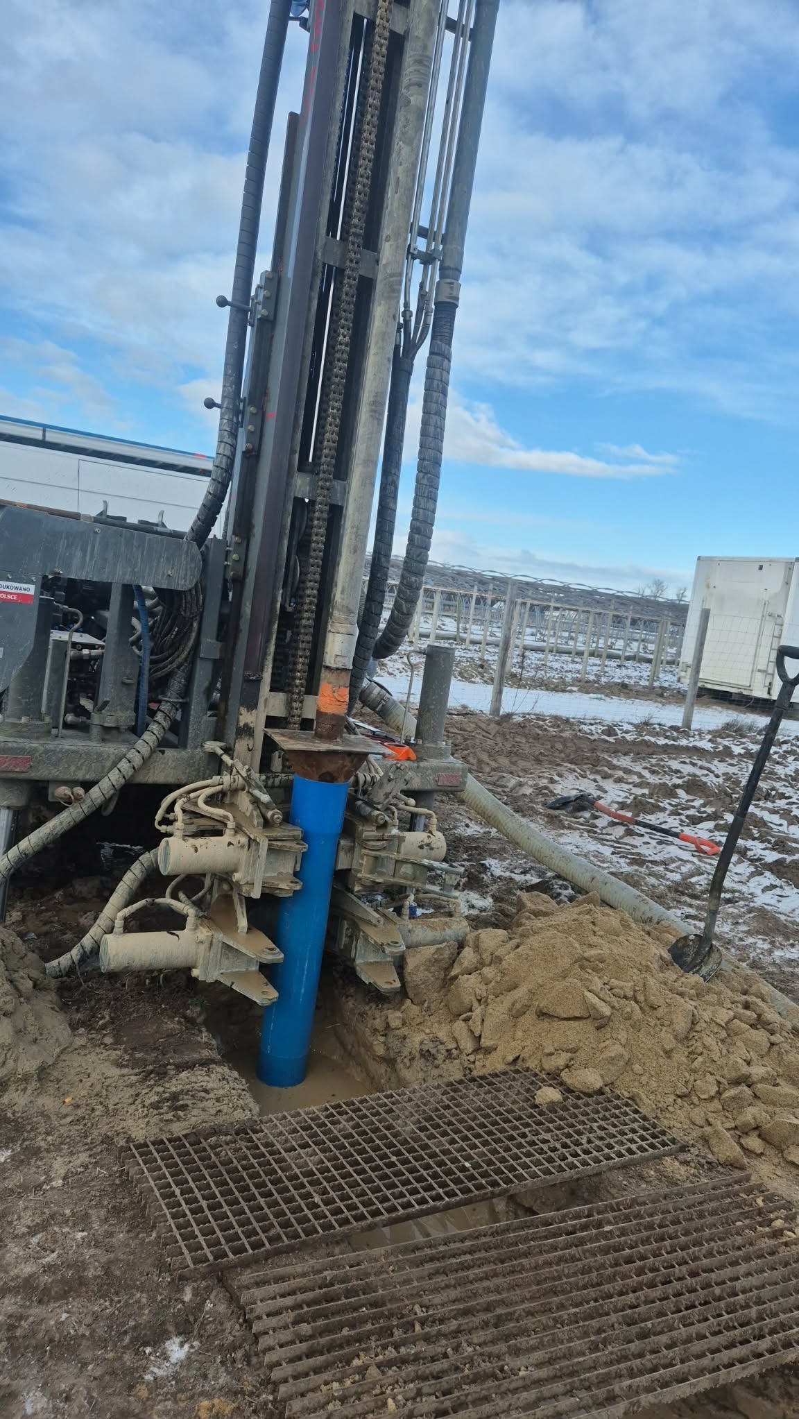 Wiertnica w trakcie pracy przy studni głębinowej w Skórzcu. Widoczna niebieska rura w ziemi, metalowa krata i sprzęt wiertniczy na tle pochmurnego nieba.