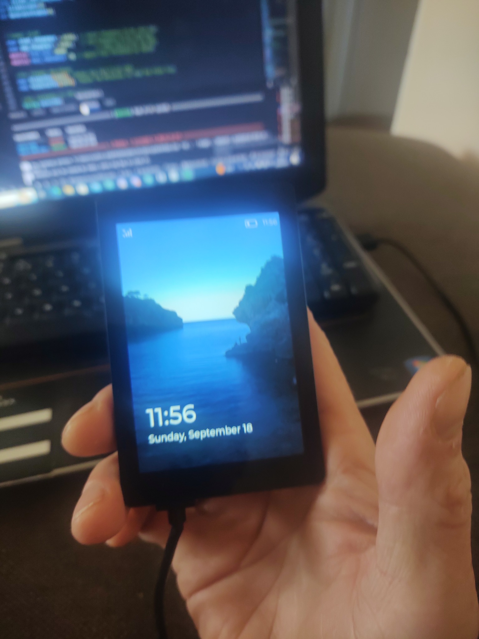 Dłoń trzyma mały, czarny ekran z wyświetloną datą i godziną oraz obrazem zatoki; w tle rozmazany ekran laptopa z kodem.