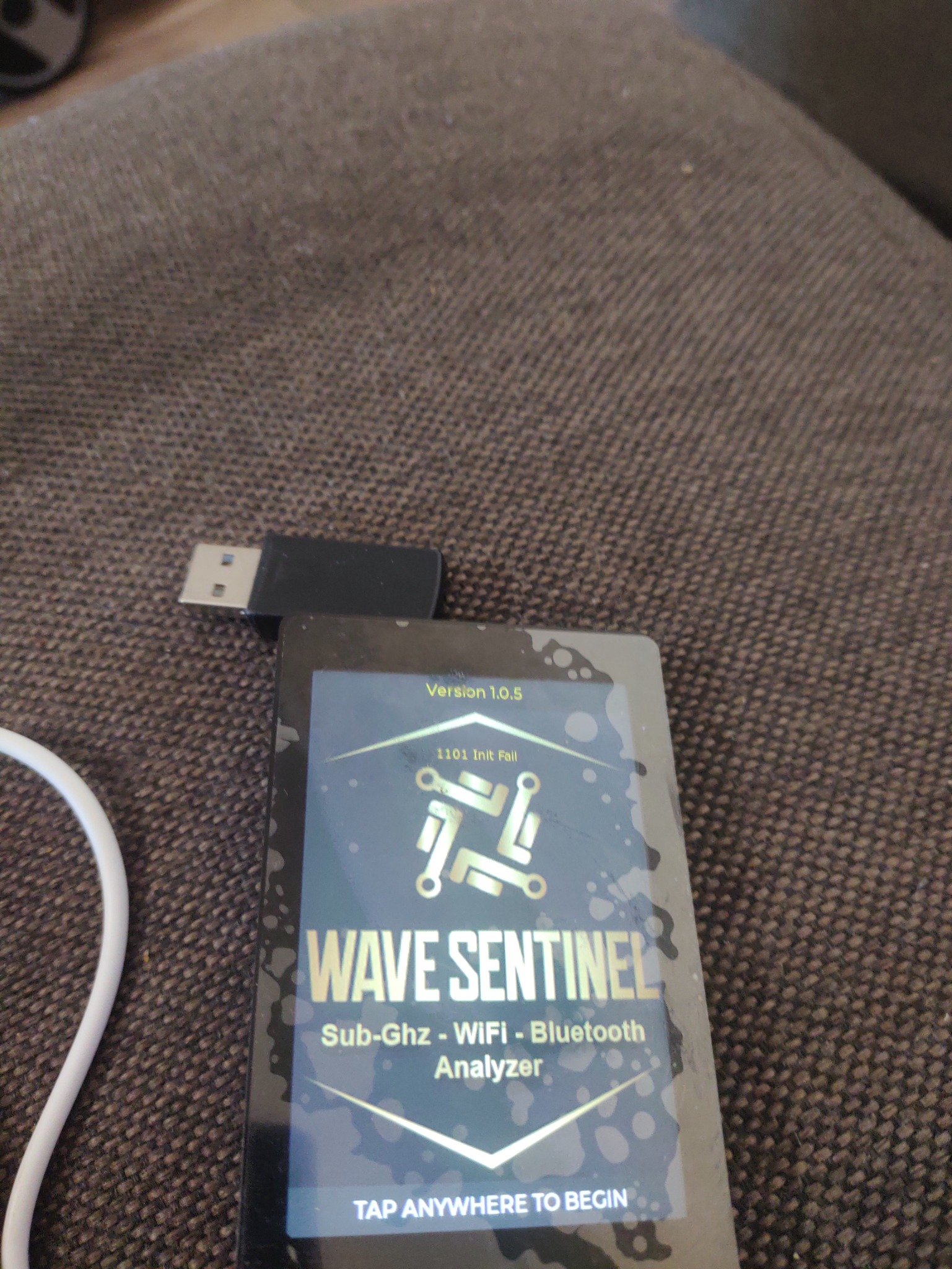 Urządzenie Wave Sentinel, analizator Sub-Ghz, WiFi i Bluetooth, z podłączonym kablem USB, leżące na brązowej, teksturowanej powierzchni. Na ekranie urządzenia widoczny napis '1101 Init Fail'.