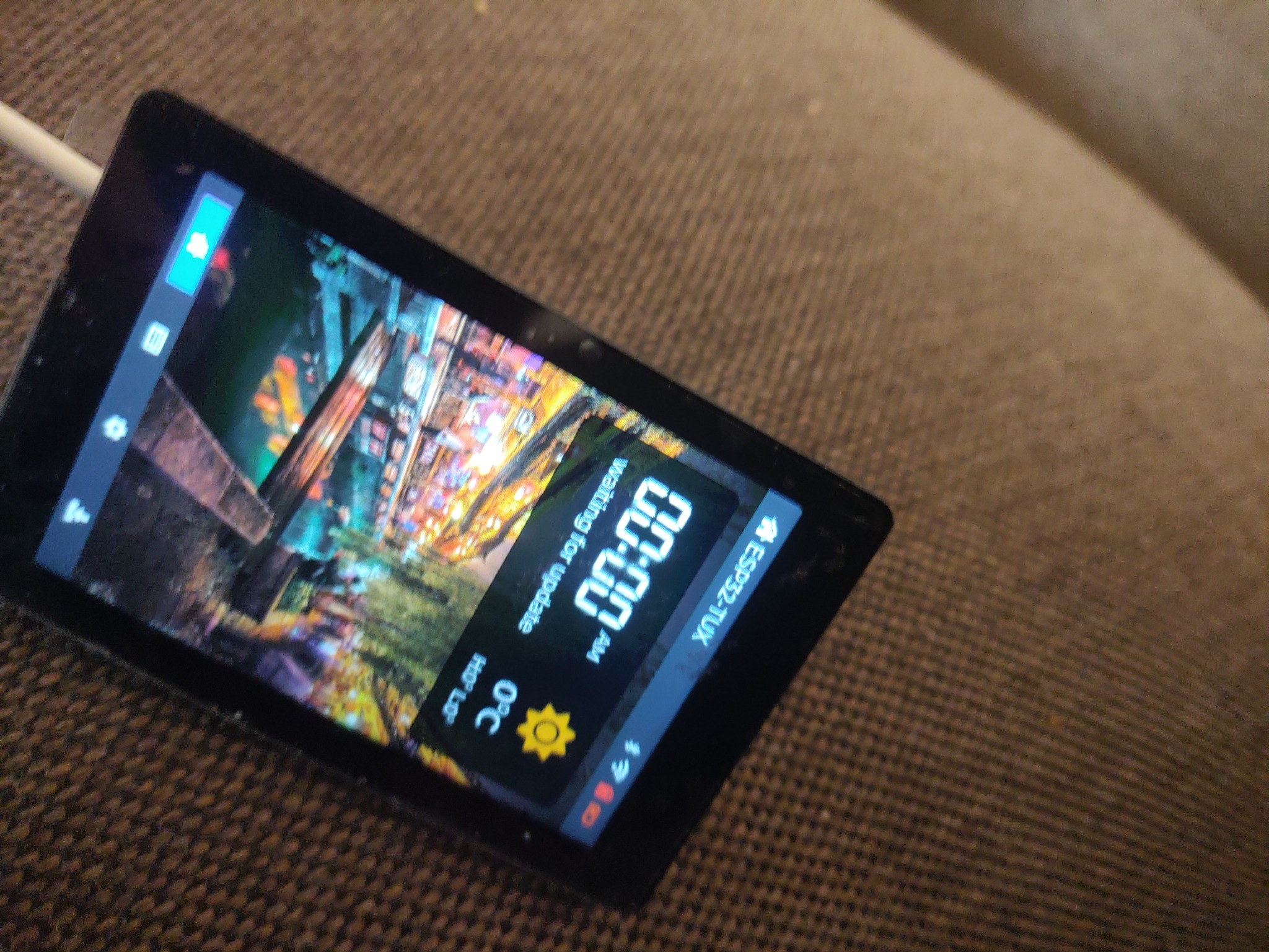 Ekran dotykowy urządzenia z wyświetlanym interfejsem użytkownika, pokazującym czas (00:00), temperaturę 0°C i status aktualizacji, umieszczony na brązowym tle.