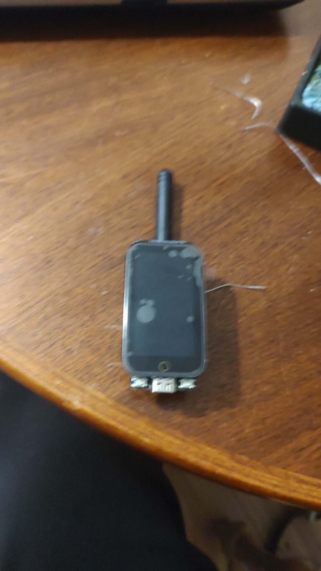 Niewielki, czarny, dotykowy moduł z anteną i portem USB leżący na drewnianym stole. Ekran modułu jest lekko zabrudzony, a obudowa porysowana. Widoczne są elementy elektroniczne na spodzie urządzenia.