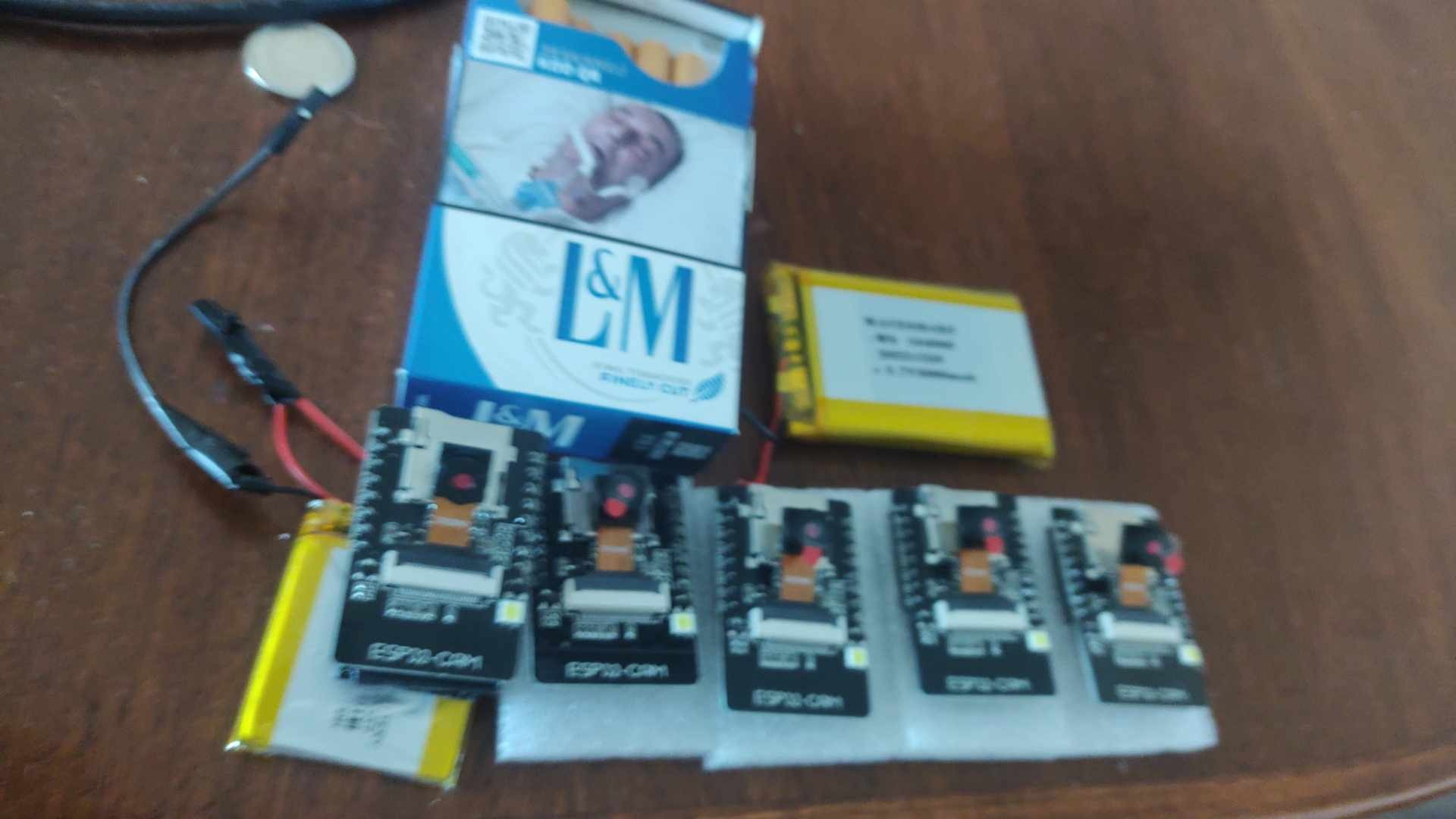 Cztery moduły ESP32-CAM z podłączonymi bateriami i przewodami, leżące na drewnianym stole obok paczki papierosów L&M i dodatkowej baterii.