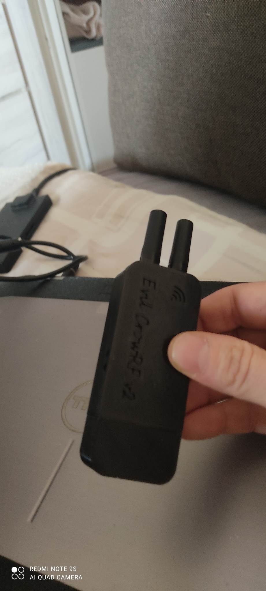 Czarny moduł z dwoma antenkami, oznaczony napisem 'End CrownRF v2', trzymany w dłoni nad laptopem Dell, w tle fragment sofy.